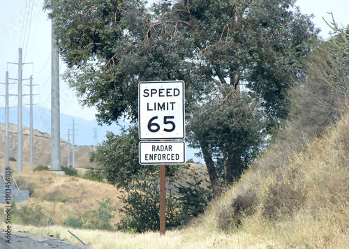 65 miles per hour speed limit sign. Radar enforced sign. On side of freeway in Southern California, trees and power lines in background