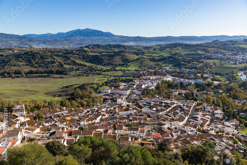 Aerial panoramic view of historic white town with tiled rooftops, located among green hills, farmland, and natural landscape. Cadiz province, Spain.