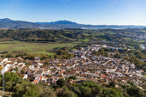 Aerial panoramic view of historic white town with tiled rooftops, located among green hills, farmland, and natural landscape. Cadiz province, Spain.