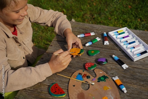 A child decorating flat pebbles with acrylic paint, depicting fresh produce like apples, carrots, and tomatoes.