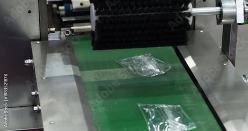 Small plastic sachets pass along conveyor under cleaning brush in packaging machine. Automated production stage with industrial equipment used in modern manufacturing process.