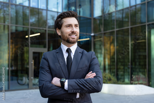 Confident businessman in a dark suit with crossed arms standing outdoors in front of a modern glass office building, smiling and looking away, representing success and professionalism