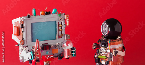 Computer robot with blue screen holding bulb and metal clamp looking at second assistant robot with spherical black head, red background. Tech device interaction concept, gadget collaboration poster