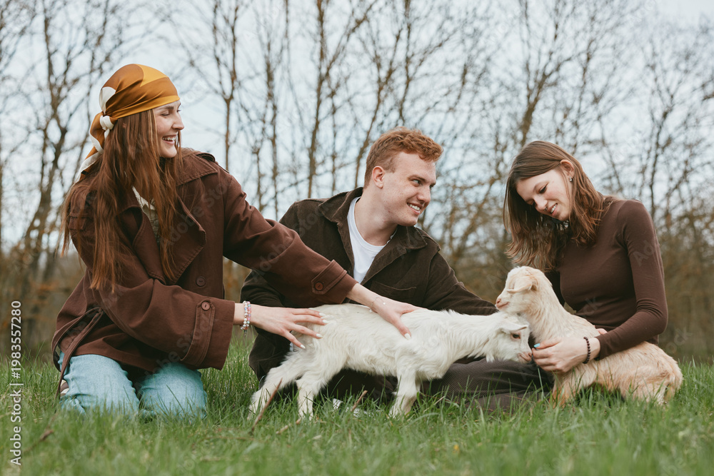 Fototapeta premium A joyful trio outdoors with a playful goat kid, as friends gently pat the animals. Casual lifestyle scene highlighting bonding, care, and rural simplicity.