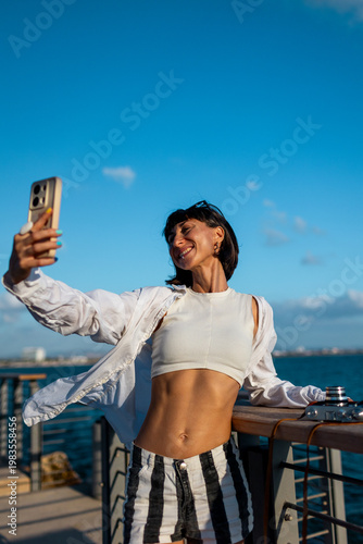 Attractive smiling tourist taking a selfie on her mobile phone against the backdrop of the Mediterranean Sea. The girl enjoys her vacation and shoots content for social media.