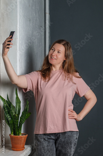 Pretty young woman with red hair in a light T-shirt and gray jeans stands against a gray wall, smiling and posing with a mobile phone. A flower on the window. Selective focus