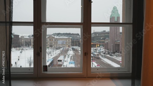 A serene view of a snowy urban landscape captured through a stylish window frame. Buildings and streets are blanketed in white, showcasing a peaceful winter scene amidst city life.
