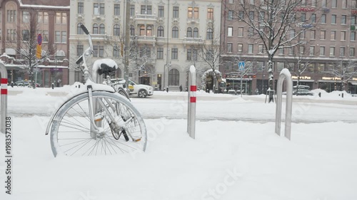 A bicycle stands in a snowy urban setting, surrounded by buildings and trees. The serene winter scene captures the beauty of a city blanketed in snow, highlighting the contrast between the vibrant urb
