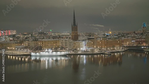 A stunning nighttime cityscape showcasing a waterfront area, with illuminated buildings, a prominent spire, and reflections dancing on the calm water.