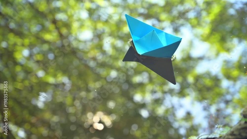 Conceptual video of a small blue paper boat slowly drifting on calm water, reflecting a rich canopy of leafy trees. Peaceful and poetic atmosphere representing nature, childhood imagination, serenity 