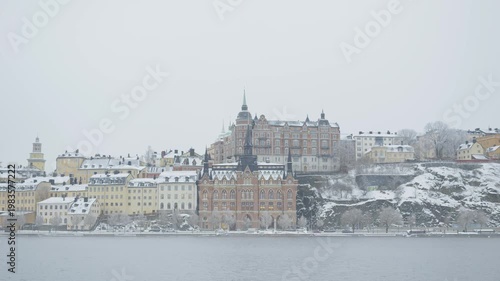 A serene winter view of a snow-covered city, showcasing a mix of historic architecture by the riverside. The scene evokes a peaceful, tranquil atmosphere, perfect for winter lovers and urban explorers
