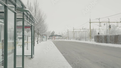 A serene winter scene featuring a snow-covered bus station, an empty road, and bare trees. The atmosphere evokes calm and solitude, perfect for winter photography.