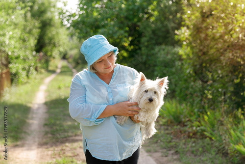 A middle-aged woman in a blue sun hat and light shirt walking along a rural path, tenderly carrying her white West Highland Terrier in her arms on a sunny day.