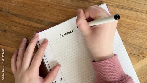 Close up of hands writing “summer rules” on lined notebook paper, casual planning and seasonal concept, natural light overhead shot on wooden table, 4K video