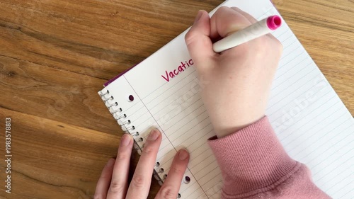 Close up of hands writing “vacation ideas” on lined notebook paper, travel planning and summer trip concept, natural light overhead shot on wooden table, 4K video