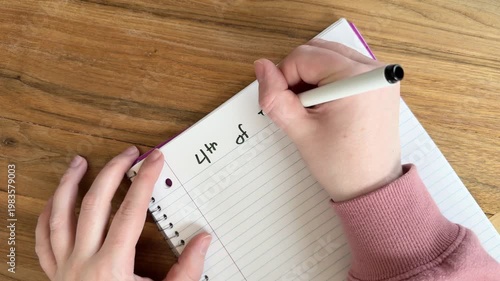 Close up of hands writing “4th of July plans” on lined notebook paper, summer holiday planning concept for Independence Day, natural light overhead shot on wooden table, 4K video