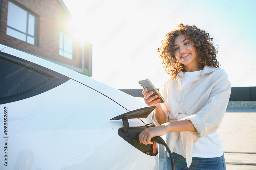Fototapeta premium Woman charging electric car at home using mobile phone app