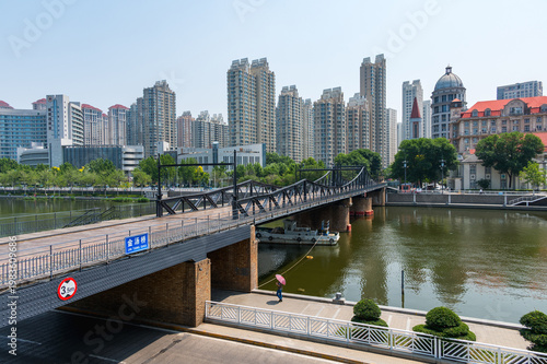 A historic steel bridge spans the Hai River in Tianjin, dense urban landscape of modern apartments and European-style architecture under a blue sky. China, 22 May 2025