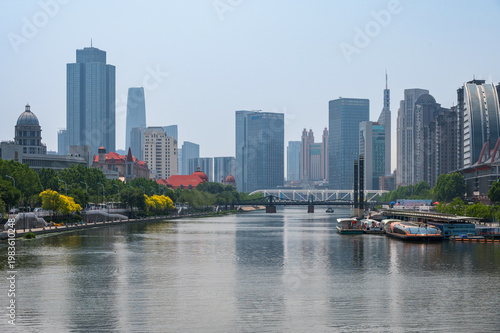 The Hai River flows through the heart of Tianjin, flanked by a mix of modern skyscrapers and green riverside promenades reflecting in the calm water. China, 22 May 2025