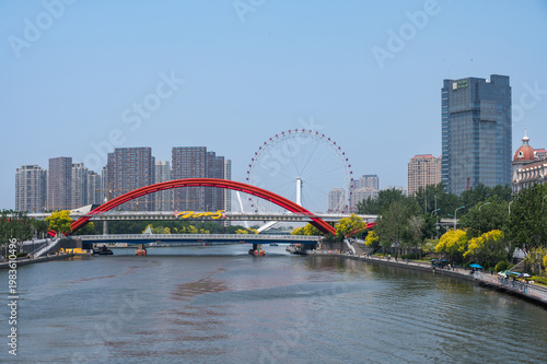 The Hai River flows through the modern cityscape of Tianjin, leading the eye toward an iconic red arched bridge and the Tianjin Eye Ferris wheel. China, 22 May 2025