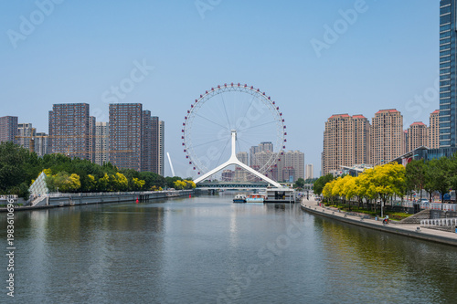 The iconic Tianjin Eye Ferris wheel towers over a city road and modern high-rise apartments under a clear blue sky. Tianjin, China - 22 May 2025