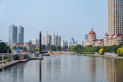 A picturesque view of the Hai River in Tianjin featuring a blend of classical European-style architecture and modern skyscrapers along the sunlit waterfront. China, 22 May 2025