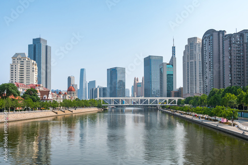 The Hai River flows through the heart of Tianjin, flanked by a mix of modern skyscrapers and green riverside promenades reflecting in the calm water. China, 22 May 2025