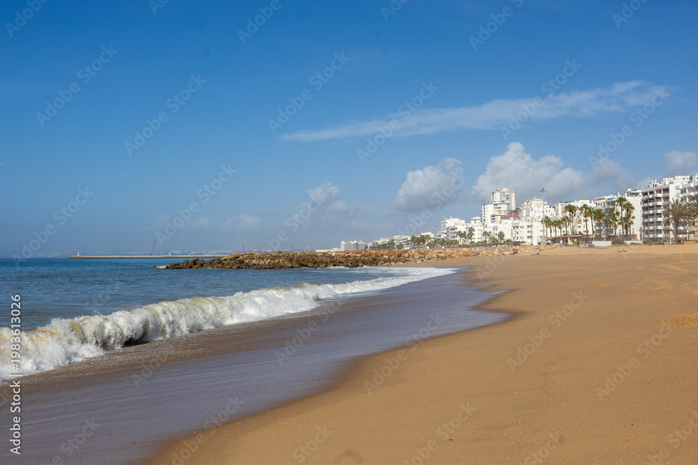Obraz premium Sandy beach with ocean waves and Faro city Algarve Portugal coastline