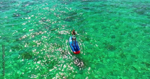 Aerial view of woman in white swimsuite standing on sup board with paddle on crystal turquoise water with coral reef on the tropical island in Indian ocean. Active water sport. Mauritius island