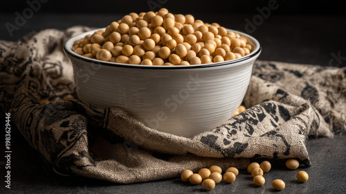 A white bowl overflowing with pale yellow soybeans sits on a wooden block with scattered soybeans on a textured surface