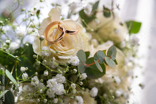 Two gold wedding rings on a bridal bouquet with white roses and gypsophila. Elegant marriage symbols and romantic floral background for engagement and wedding concepts, selective focus
