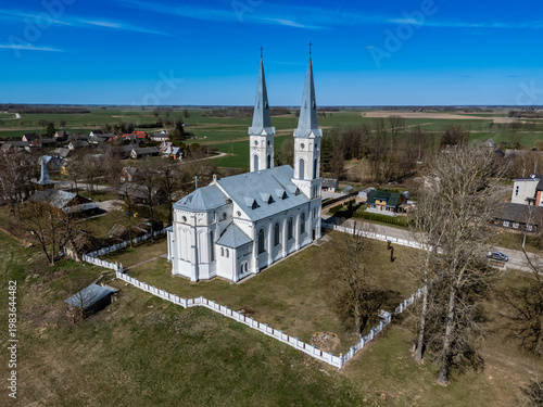 Drone aerial view of a white church with two tall towers located in a rural village surrounded by fields and houses. Scenic countryside landscape under clear blue sky with traditional European archite