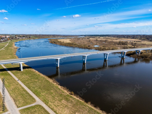 Aerial drone view of a modern bridge crossing a wide Nemunas river in a rural landscape with fields and small town in the distance under blue sky.