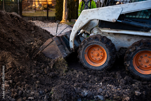 Obraz na plátně A skid steer with orange wheels bites into wet soil at dusk, bucket tilted, worker in a high visibility jacket nearby, gravel paths and conifers behind a fence