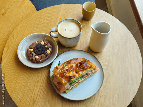 Warm wooden table holds breakfast spread. Coffee cup, small pitcher, and two plates. One plate holds quiche slice with ham and green vegetables. Other plate has chocolate cookie with nuts