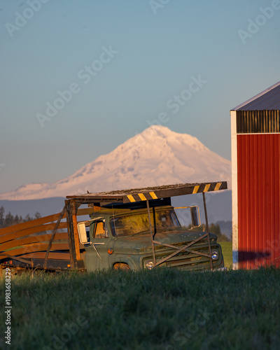 Vintage truck parked beside red barn. Snow-capped mountain rises in background. Grass field stretches in foreground. Clear blue sky above scene. No animals or plants visible