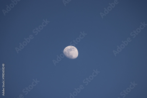 Crescent moon shines against a deep blue sky. The moon's surface shows craters and light and dark areas. No clouds or other objects are visible. The sky is clear and calm