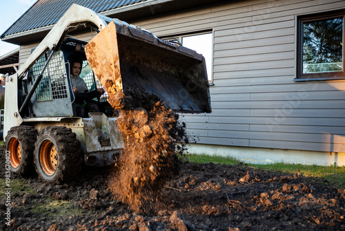 A compact skid steer loader dumps wet soil beside a modern clapboard house near dusk, operator visible, mud splatters, rutted earth, strong side light, worn tires.