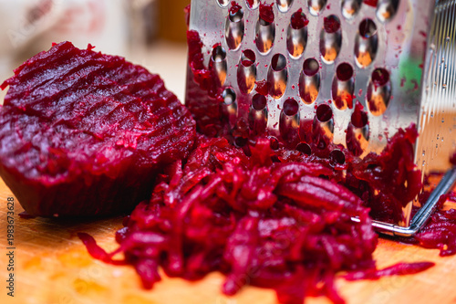 Macro of boiled red beetroot being grated on a metal grater on a wooden board. Healthy cooking process for salad or borscht. Vibrant magenta vegetable food texture