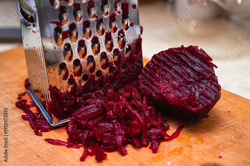 Macro of boiled red beetroot being grated on a metal grater on a wooden board. Healthy cooking process for salad or borscht. Vibrant magenta vegetable food texture