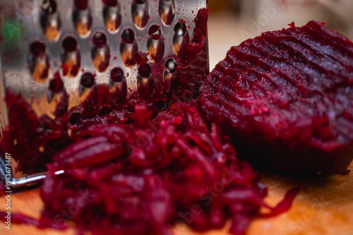 Macro of boiled red beetroot being grated on a metal grater on a wooden board. Healthy cooking process for salad or borscht. Vibrant magenta vegetable food texture