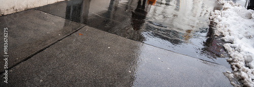A wet sidewalk reflecting surrounding buildings and pedestrians. The scene captures a chilly atmosphere with patches of snow, suggesting winter weather in an urban environment.