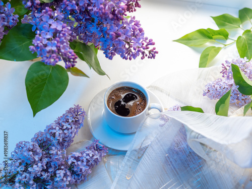 Hot coffee in a white cup on a white table with blooming purple lilac. Colorful spring greeting card