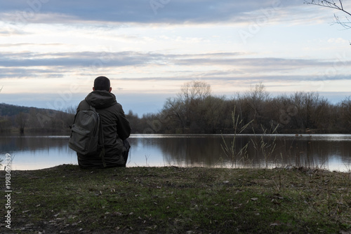 alone man with a backpack sits on the riverbank in cloudy weather. The atmosphere of depression and loneliness