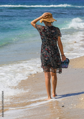 Traveler girl walking in a floral black dress on a tropical beach. Young girl traveler walking in nature. Traveler girl in a dress walking on the beach. Girl traveler walking on the beach. Summer trop