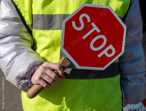 Fotografie Person wearing neon lime yellow high visibility safety vest and silver winter jacket holding wooden handle of octagonal red traffic stop paddle to regulate vehicle movement