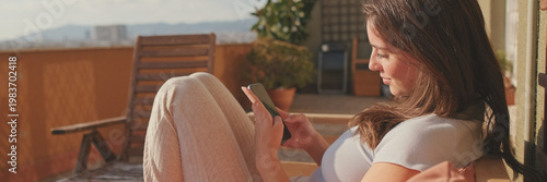 Young woman using mobile phone while sitting on balcony at home. Panoramic