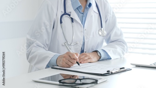Fotografía Female doctor documents patient information while working with digital devices at minimalist white workstation in medical office