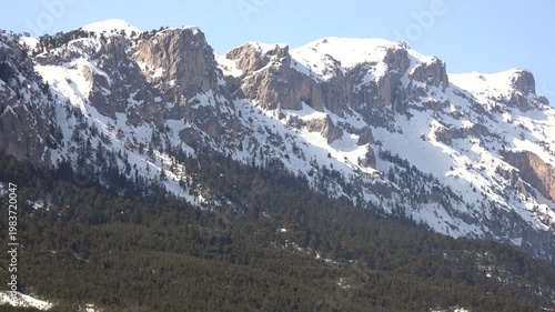 Snowy rocky Alpine mountain range rises above dark forested slopes across vast European highlands. Majestic winter peaks dominate natural landscape spanning central europe with rugged stone relief.