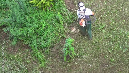 Gardener trimming grass with string trimmer on lush green outdoor garden slope area. Worker maintains vegetation using cutting equipment while walking along uneven terrain.